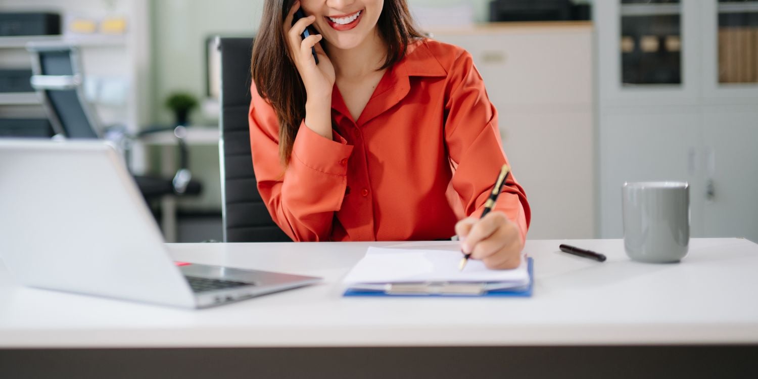 Smiling Asian woman speaks on phone while working at desk with l